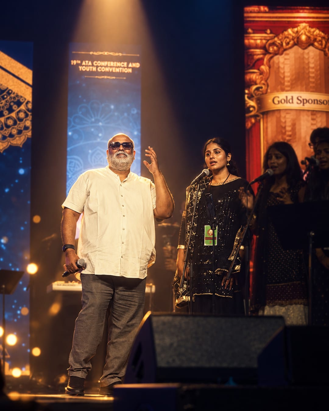 Hasitha singing at the microphone under a spotlight at the 19th ATA Conference, M. M. Keeravani speaking to the hall beside her.