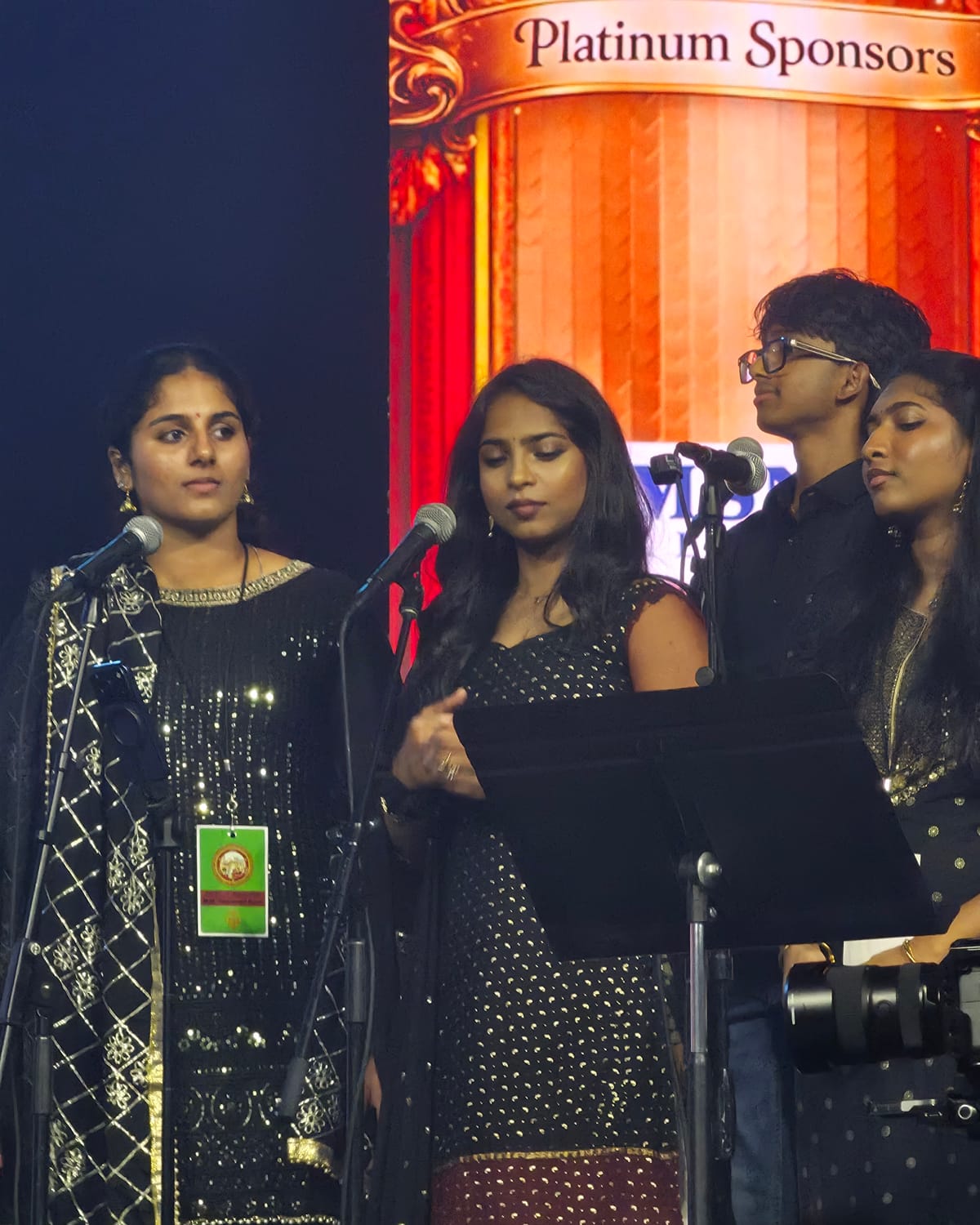 Hasitha at the microphone in a black and gold outfit, stage banner behind her.