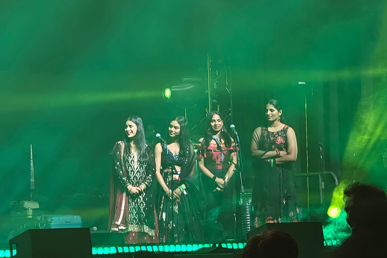 Four singers under green stage light and smoke, audience silhouetted in front.